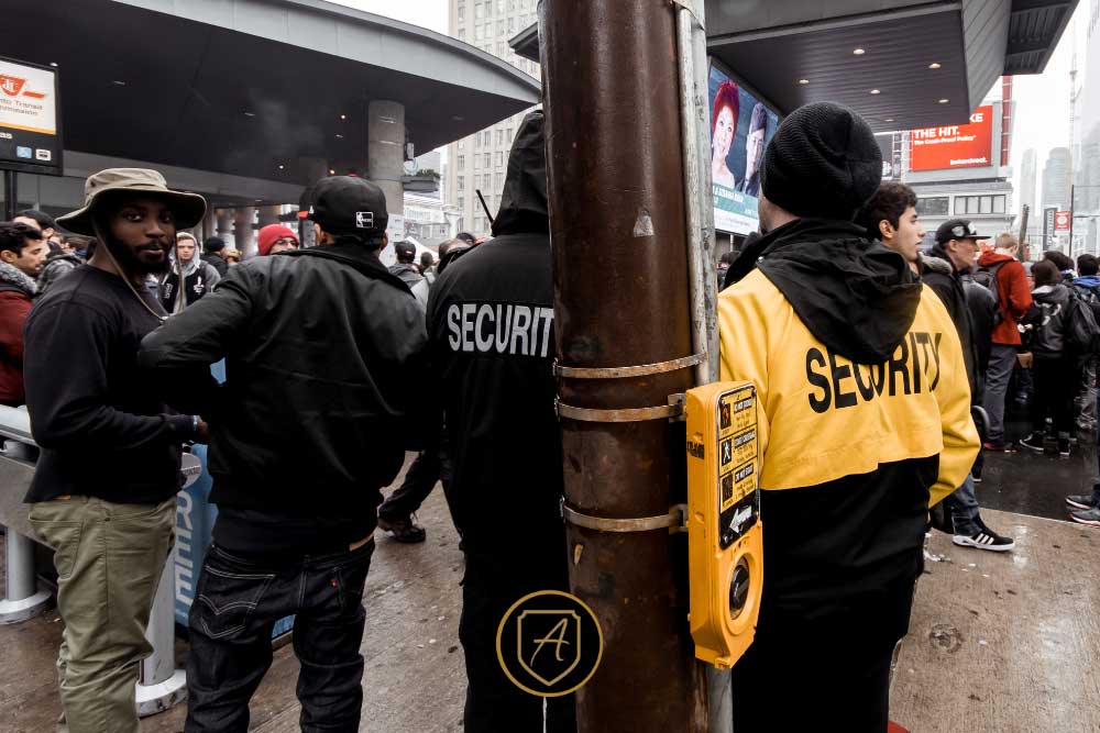 Outside a building entrance, two uniformed security guards—one in black, one in yellow—provide executive protection among a crowd, highlighting the real value of uniformed security guards for Nevada businesses. A yellow-buttoned pole stands in front. | Executive Protection & Bodyguards in Las Vegas | Alexios
