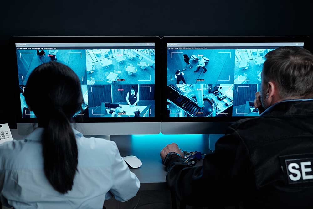Two security personnel in a dimly lit control room monitor multiple feeds on two screens, ensuring the safety of executives and high-profile clients—highlighting The-Real-Value-of-Uniformed-Security-Guards-for-Nevada-Businesses-4. | Executive Protection & Bodyguards in Las Vegas | Alexios