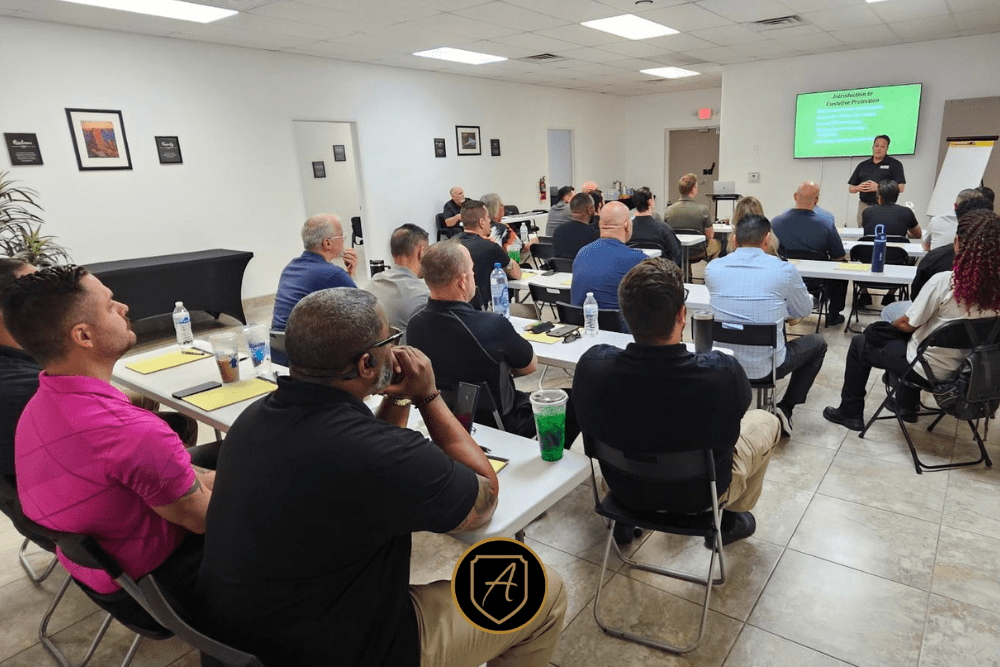 Adults sit at tables in a brightly lit classroom for the Executive Protection Training: ConOps for Protectors Course3, facing a presenter near a screen displaying travel security content. Notebooks, drinks, and water bottles are on the tables. | Executive Protection & Bodyguards in Las Vegas | Alexios