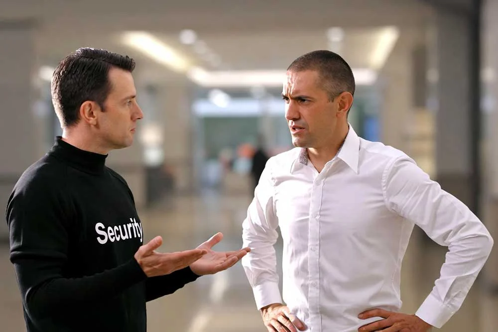 A security personnel in a black shirt labeled "Security" discusses with a man in a white shirt in a bright, modern hallway, illustrating professional security team coordination for effective event security.| Alexios Security Services | Las Vegas Executive Security