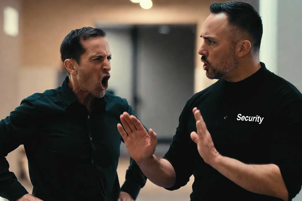 A security personnel in a black shirt labeled "Security" calmly gestures to de-escalate a situation with an upset man, exemplifying professional security services in action.| Alexios Security Services | Las Vegas Executive Security